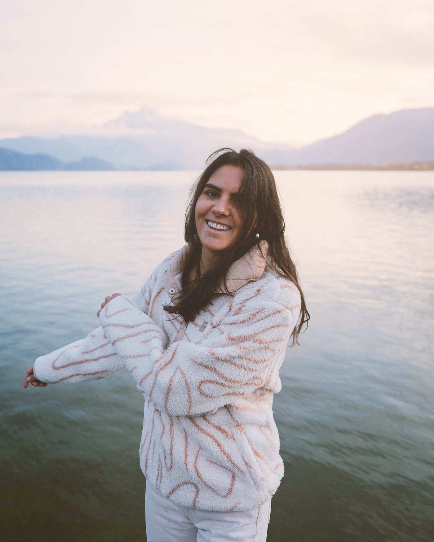 Photo of Giulia smiling at the camera, posing in front of a large lake with mountains in the back