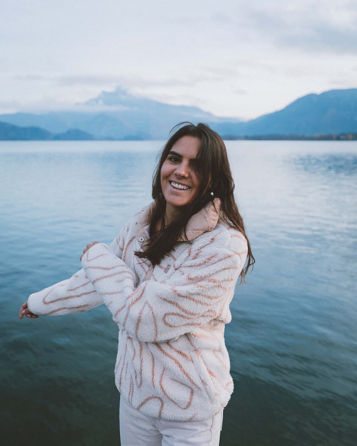 Photo of Giulia smiling at the camera, posing in front of a large lake with mountains in the back